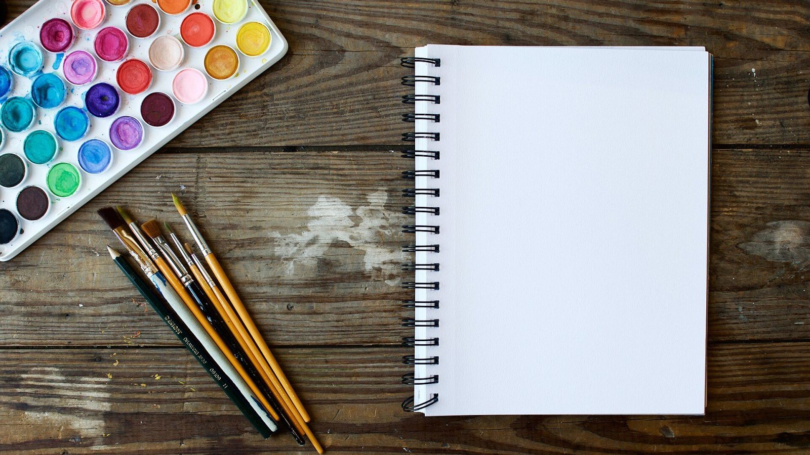 علبة ألوان مائية وفرش رسم ودفتر رسم أبيض على طاولة خشبية